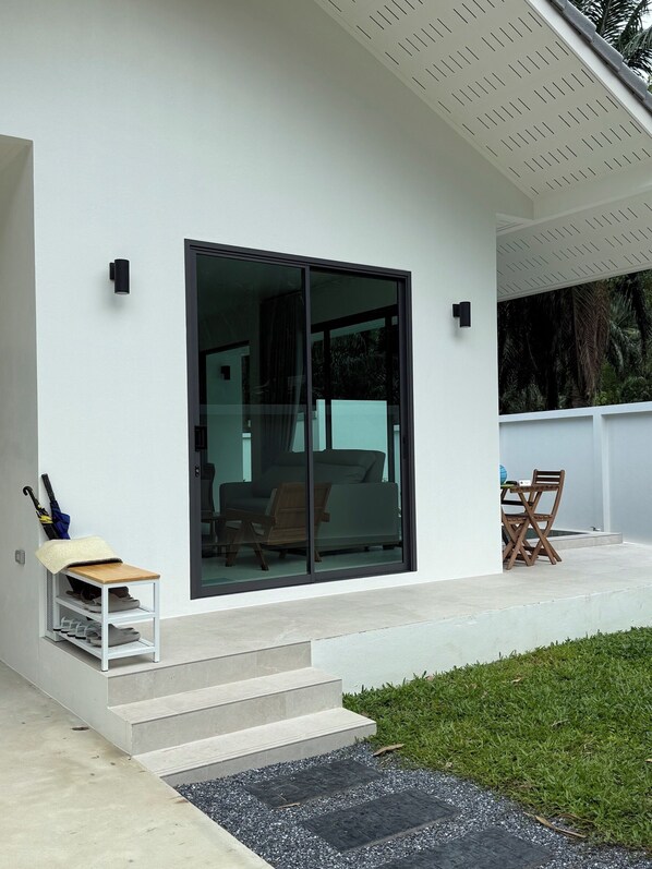 A modern house with a large sliding glass door leading to a living area, a small outdoor seating area with a table and chairs, and a covered shoe rack with an umbrella. - Passara Villas (Krabi)