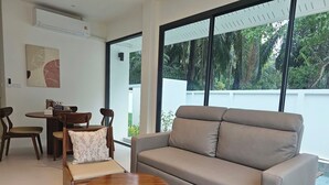 A modern living room with a sofa, chairs, a round wooden table, and a dining area with a table and chairs. - Passara Villas (Krabi)