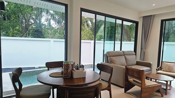 A modern living room with a round dining table, wooden chairs, a sofa, and a large window offering a view of a swimming pool and greenery. - Passara Villas (Krabi)