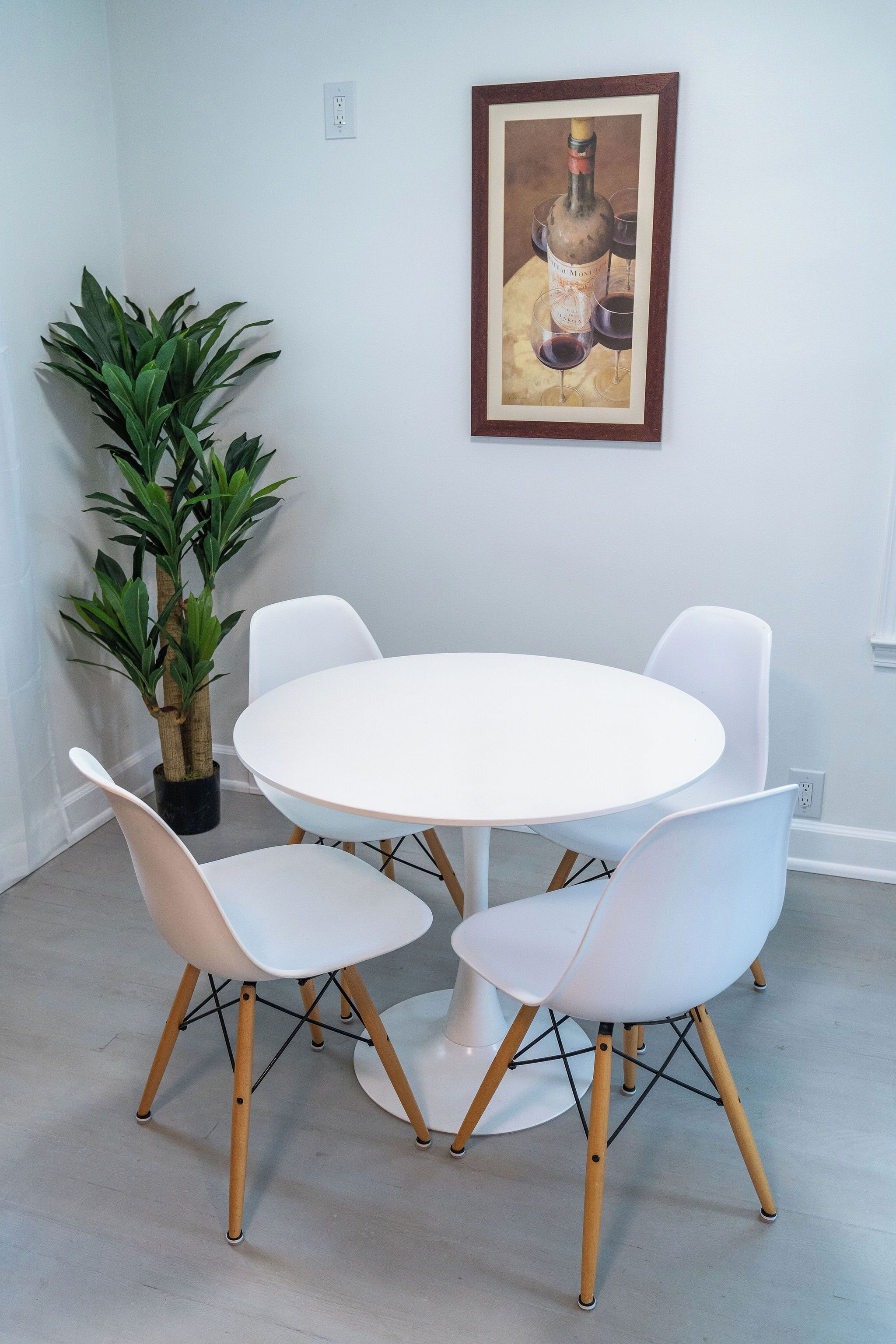 Ein Essbereich mit einem runden weißen Tisch und vier weißen Stühlen mit Holzbeinen. Eine Kübelpflanze ist links und ein gerahmtes Kunstwerk hängt über dem Tisch.
