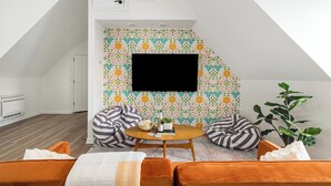 A cozy loft bedroom with a sloped ceiling, a large TV mounted on the wall, a round wooden coffee table, and a comfortable seating area with orange and brown cushions. - Family FUN! Luxury Touches, Mini Golf, Firepit, Fully Loaded (Thompson's Station)