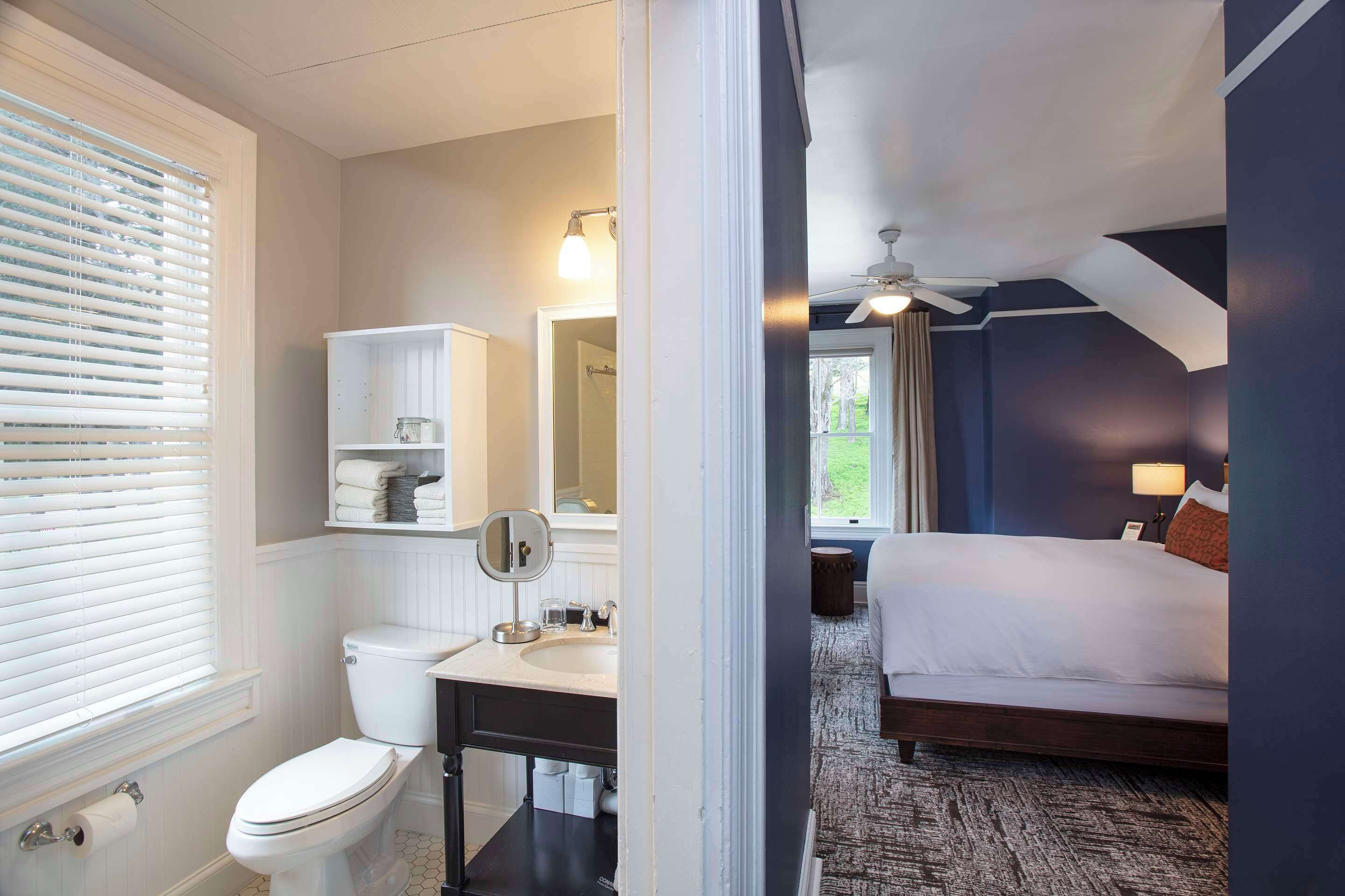 A hotel room with a bathroom, a bed, a ceiling fan, and a view of the outdoors.