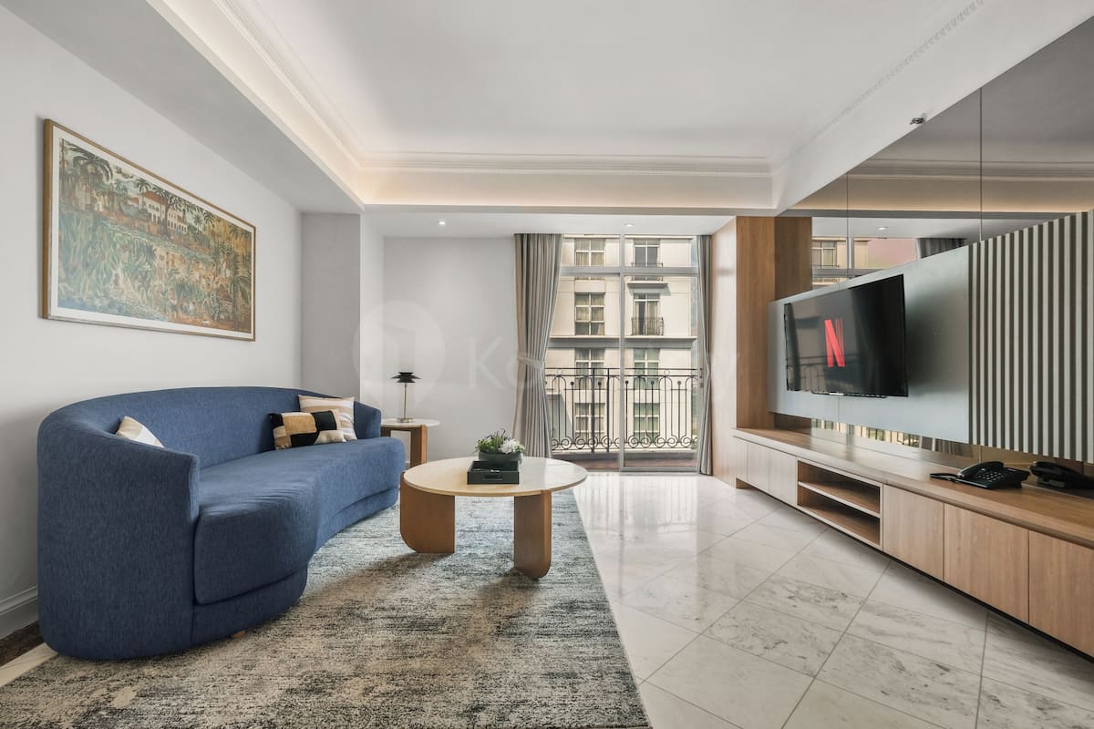 Un salon moderne avec un canapé bleu, une table basse, un téléviseur et un balcon offrant une vue sur les immeubles.
