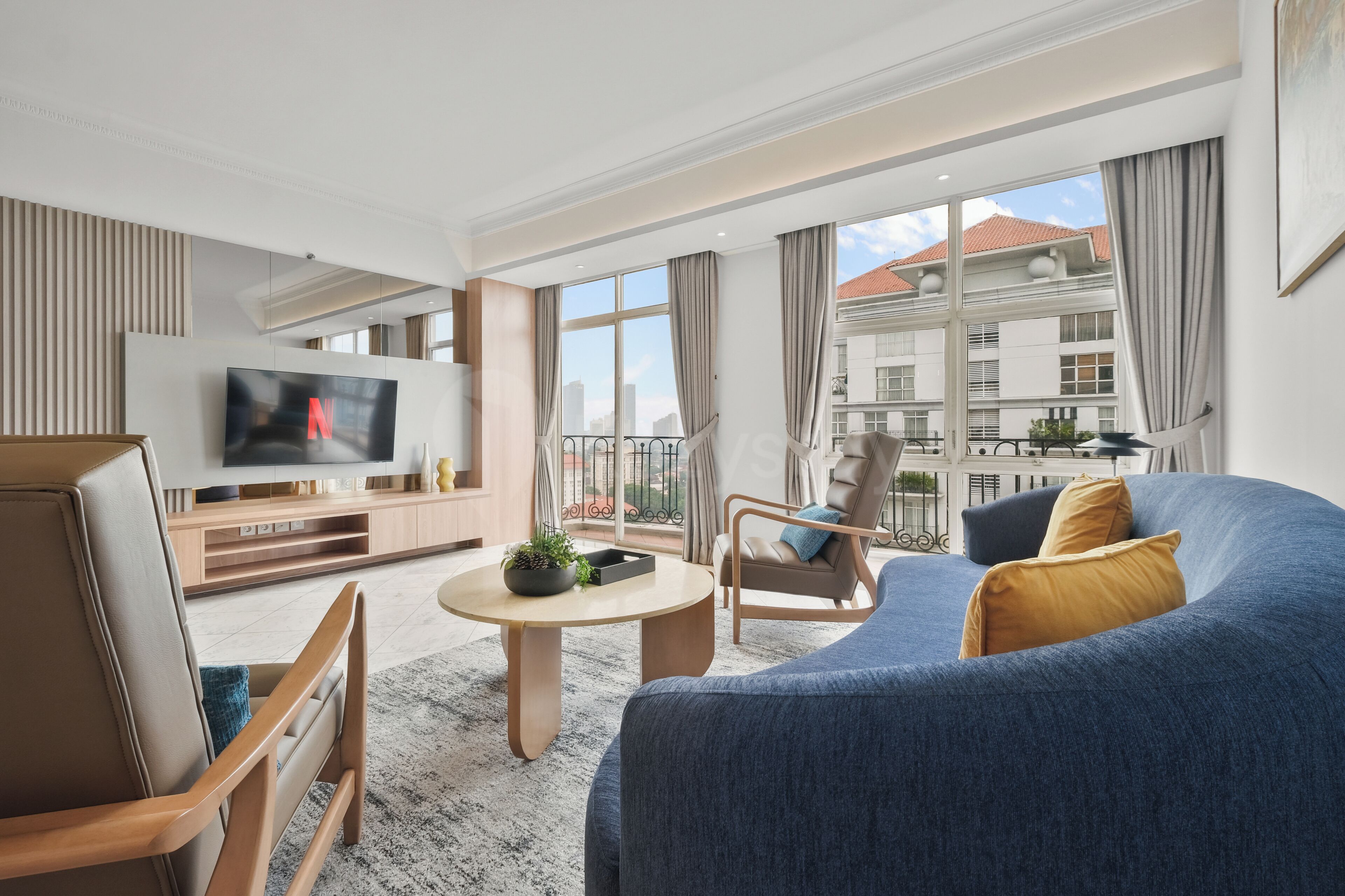 Ein modernes Wohnzimmer mit einem großen Flachbildfernseher, einem blauen Sofa, einem Holztisch und einem Balkon mit Blick auf Gebäude.
