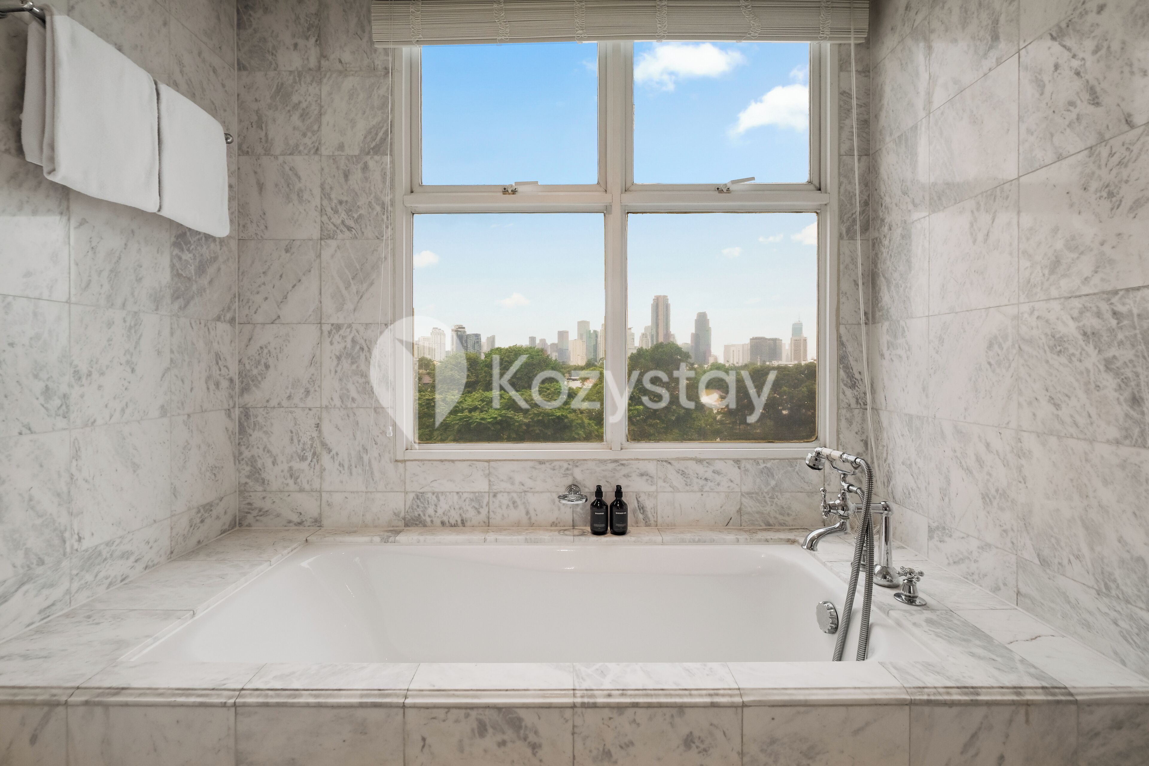 A bathroom with a large bathtub, a window with a city view, and two bottles on the edge of the tub.