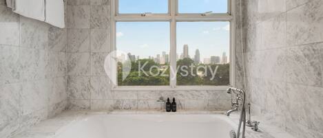 A bathroom with a large bathtub, a window with a city view, and two bottles on the edge of the tub.