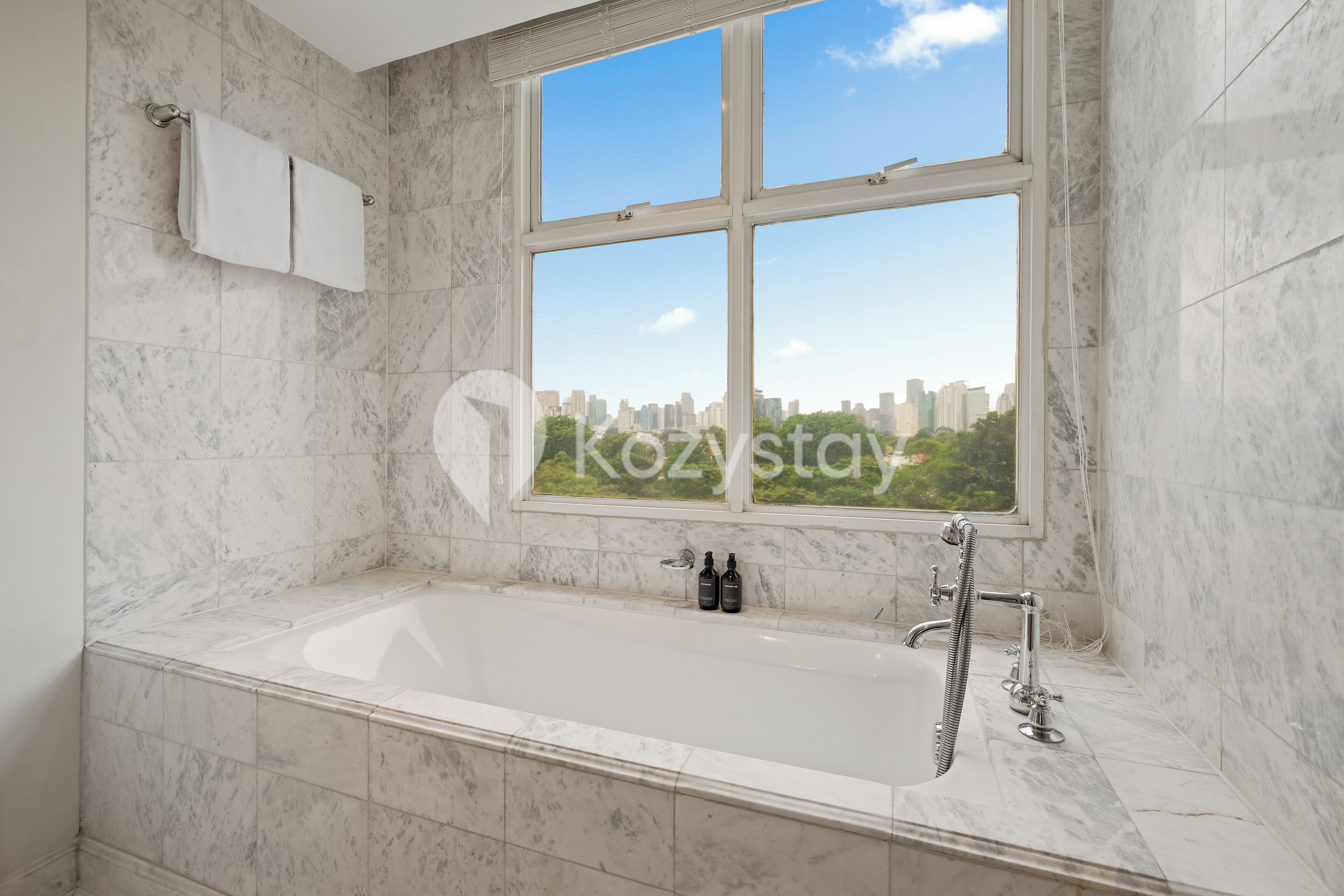 A modern bathroom with a large bathtub, a window with a city view, and marble tiles.