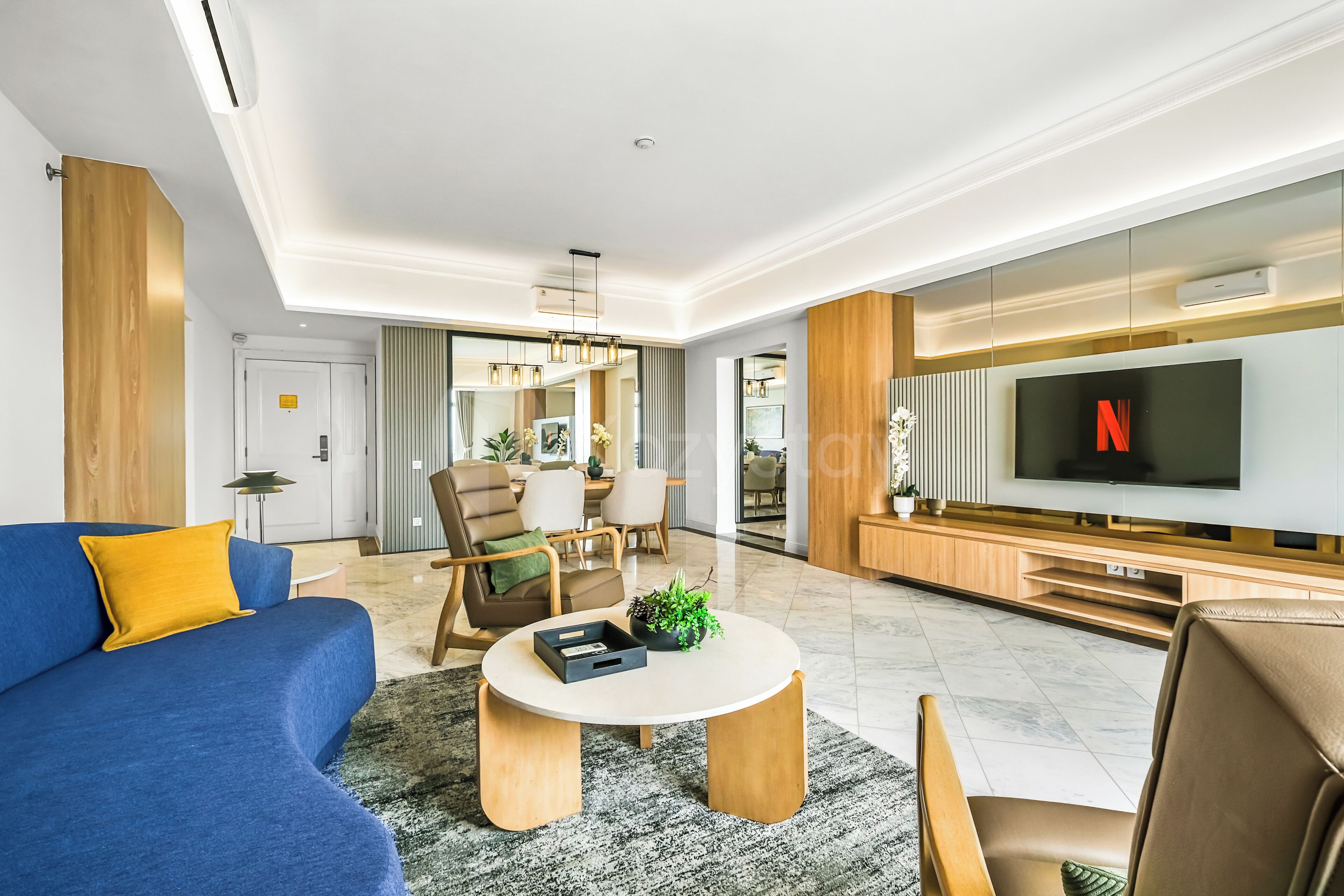 A modern living room with a blue sofa, a round coffee table, a TV, and a dining area in the background.