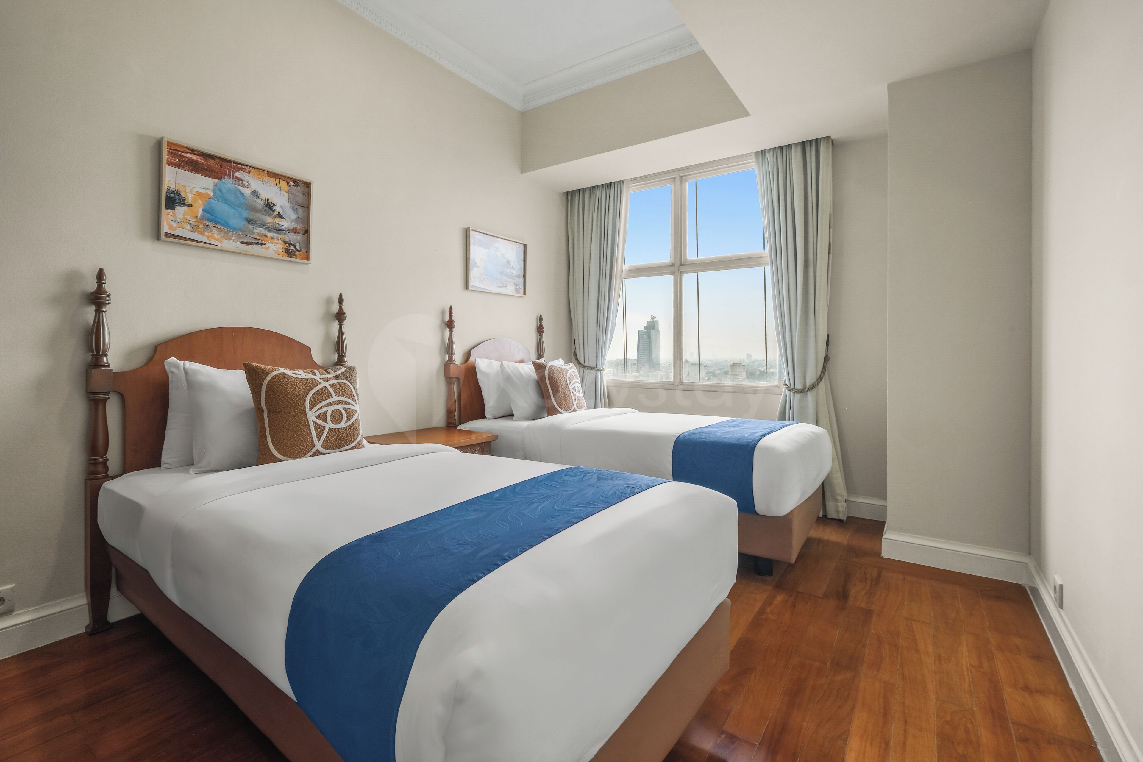 A hotel room with two beds, a large window with curtains, a painting on the wall, and a view of the city.