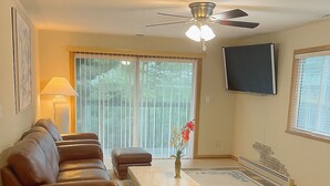 A living room with a brown leather sofa, a wooden coffee table, a floor lamp, a TV mounted on the wall, and a large window with blinds.