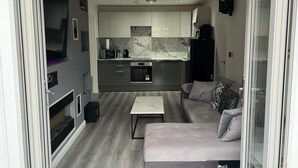A modern kitchen with a marble backsplash, a built-in oven, and a black refrigerator. There is a grey sofa with patterned cushions and a white coffee table in the living area.