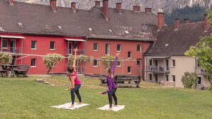 Dos personas practicando yoga al aire libre frente a un edificio rojo con montañas al fondo.