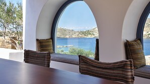 A room with a view of the sea, featuring a wooden table and two cushioned seating areas with patterned cushions.