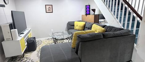 A modern living room with a grey sofa, a glass coffee table, and a staircase with white railings.