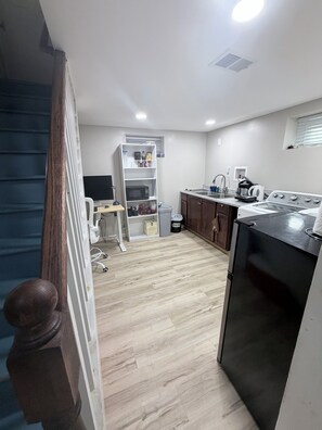 A compact kitchen with a microwave, stove, and refrigerator. A white shelf unit with various items is visible in the background.
