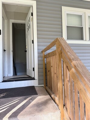 A house entrance with a wooden front door, a small porch with a wooden railing, and a window with blinds.