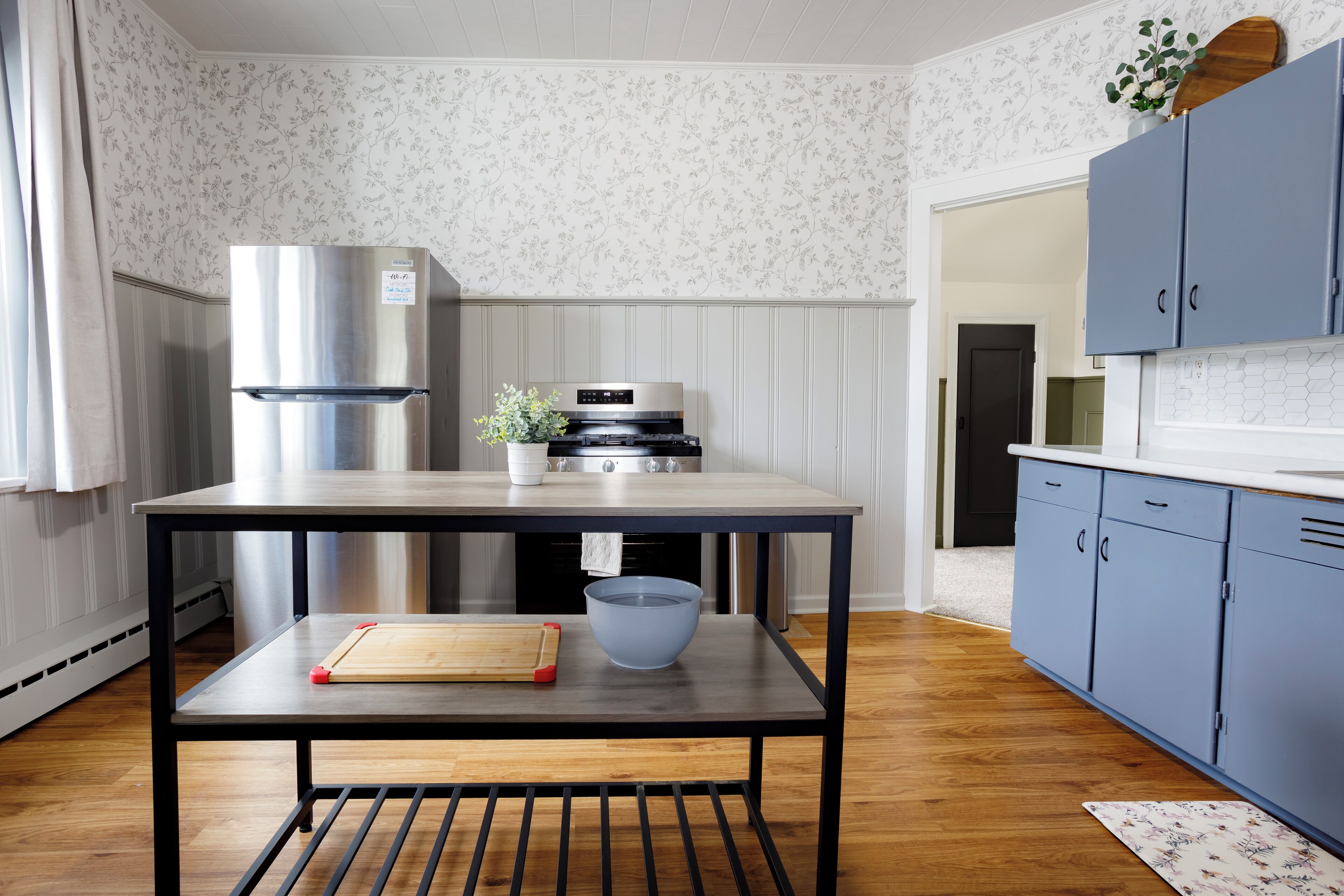 Una cocina con un refrigerador de acero inoxidable, una mesa de madera con tabla de cortar y un tazón, y armarios azules.