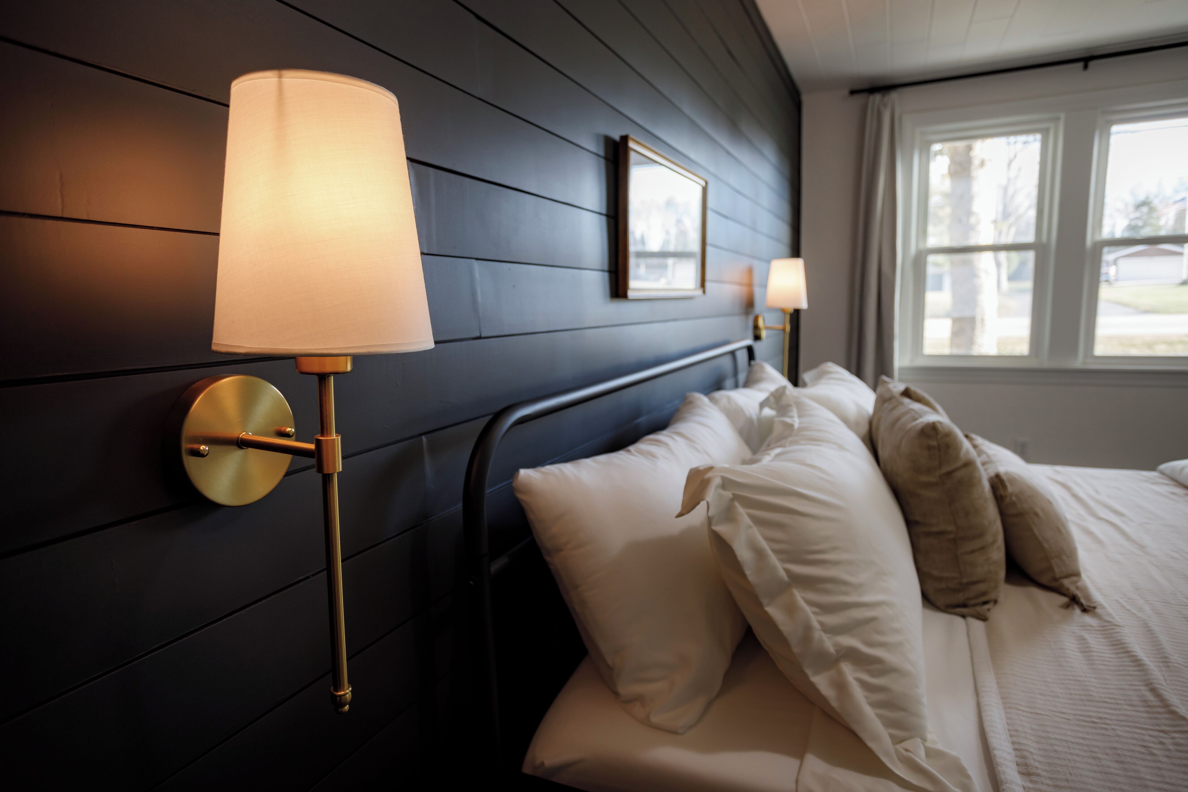 Un lit bien fait, avec des oreillers blancs et beiges, une tête de lit en bois foncé et deux lampes fixées au mur, dotées d’abat-jours blancs.