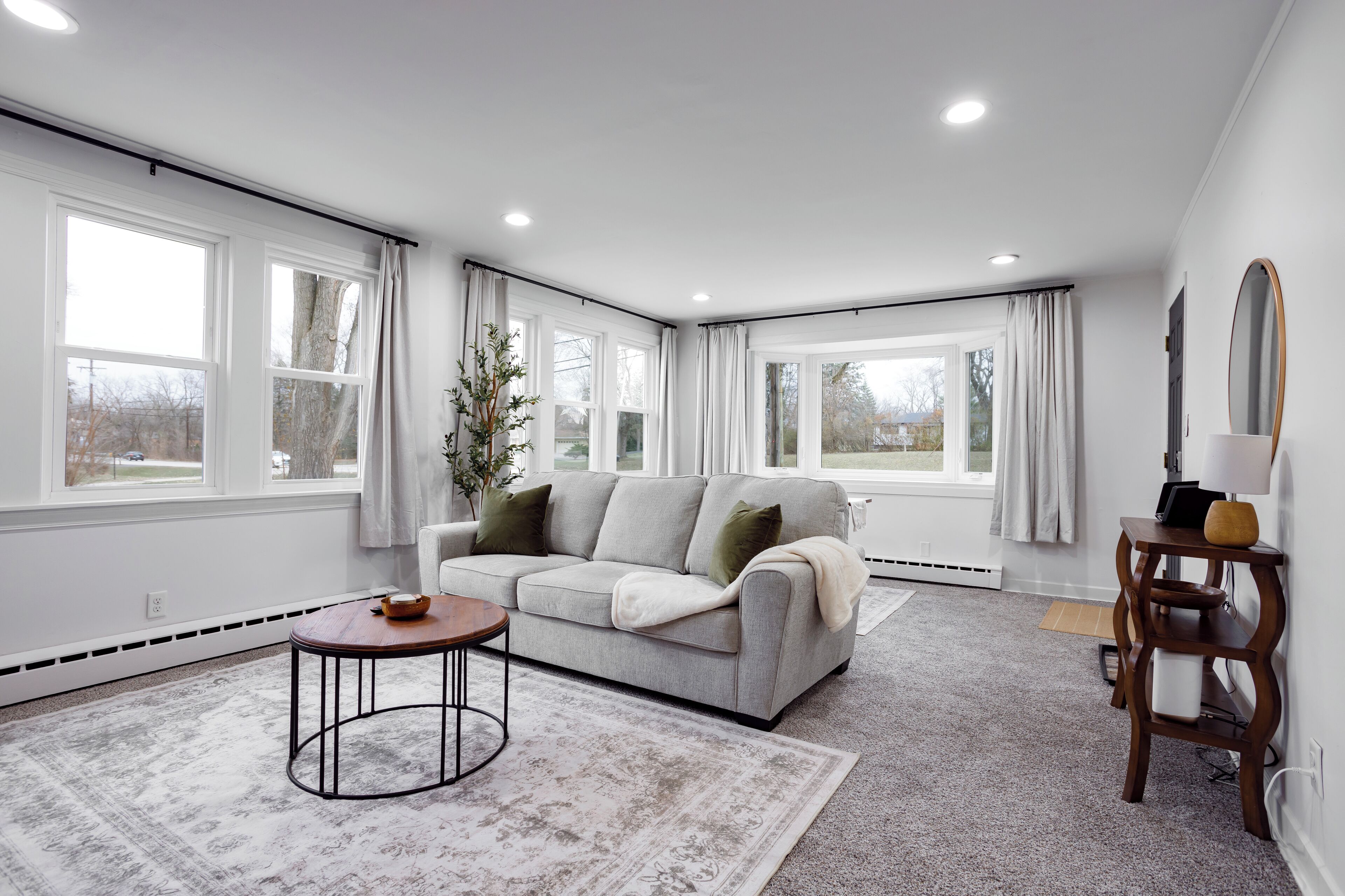 Een moderne woonkamer met een grijze bank, een ronde salontafel en een groot raam met gordijnen.