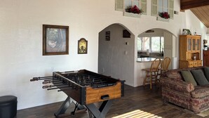 A cozy living room with a foosball table, wooden furniture, and a kitchen area visible through an archway.