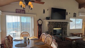 A dining area with a wooden table and chairs, a stone fireplace, a television mounted above the fireplace, and a view of the outdoors through large windows.