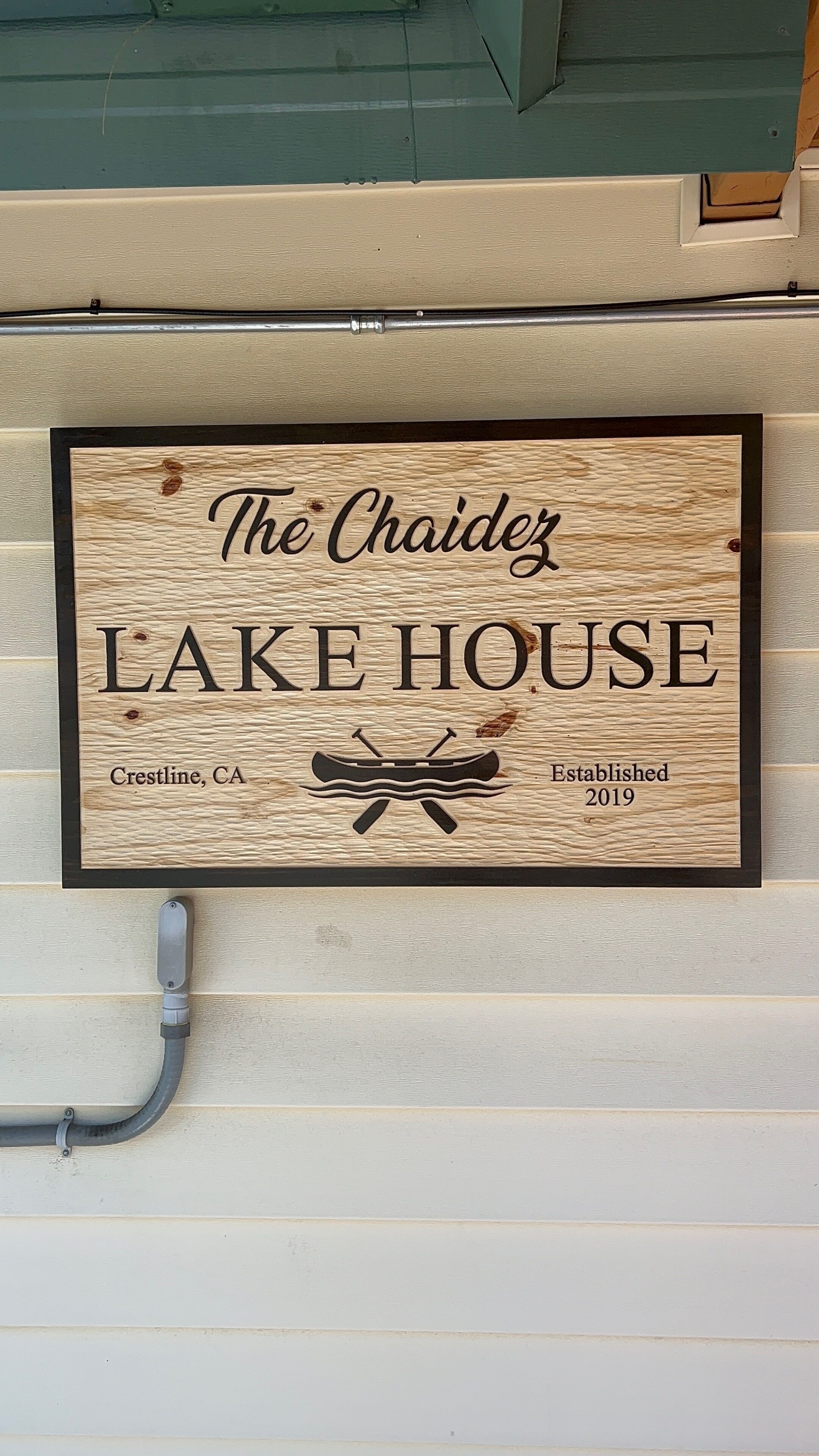 Un letrero de madera con el texto 'The Chaidez LAKE HOUSE' y un gráfico de canoa, indicando un establecimiento frente al lago.