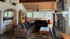 A cozy living room with a fireplace, wooden ceiling beams, a dining area, and a kitchen with a large window.