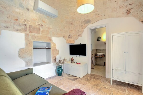 A living room with a stone wall, a flat-screen TV, a sofa, a cabinet, and a bathroom visible in the background. - Dimora Tra Gli Ulivi With Private Jacuzzi, Ostuni, Italy (Ostuni)