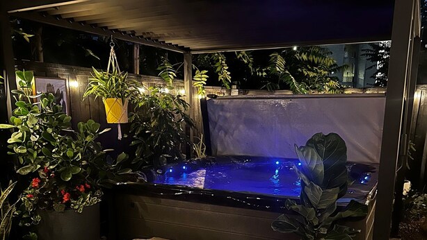 Un espace avec un bain à remous, accessible par des marches en pierre, entouré de plantes en pot et agrémenté d’une pergola illuminée de guirlandes lumineuses.