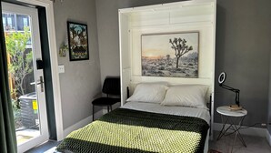 Une chambre à coucher avec un lit, un tableau encadré représentant un paysage désertique, une petite table de chevet avec une lampe, et une vue sur l’extérieur par une porte.
