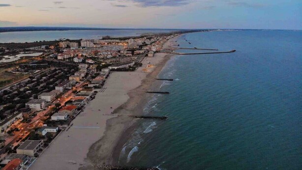 An aerial view of a coastal city with a long beachfront, a pier extending into the sea, and residential buildings lining the shore.