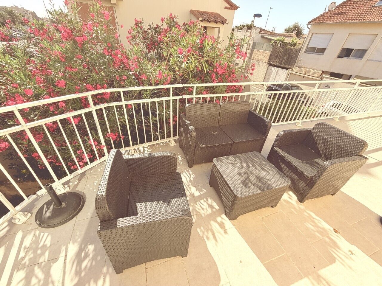 Un balcon agrémenté de meubles en osier, d’une balustrade en métal blanc et offrant une vue sur des plantes à fleurs roses.