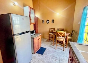 Cocina compacta con refrigerador, microondas, armarios de madera, una mesa de comedor con sillas y un ventanal con cortinas.