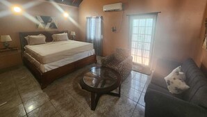 A bedroom with a bed, a chair, a sofa, a round table, an air conditioning unit, and a window.