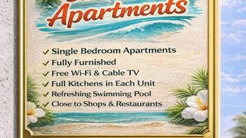 A framed sign for Crooks Apartments with a beach scene, listing amenities like single bedroom apartments, fully furnished rooms, free Wi-Fi, cable TV, full kitchens, a swimming pool, and proximity to shops and restaurants.