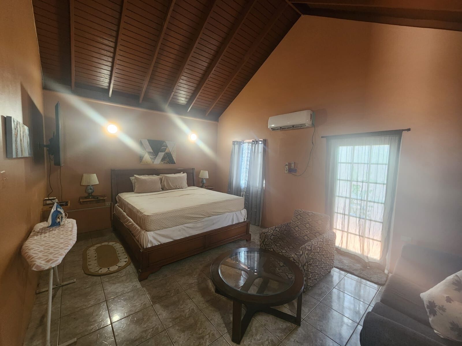 A bedroom with a bed, bedside tables, a round glass table, a sofa, a window, and an air conditioning unit.