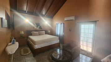 A bedroom with a bed, bedside tables, a round glass table, a sofa, a window, and an air conditioning unit.