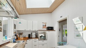 A modern kitchen with white cabinets, a built-in oven, a microwave, and a sink. There is a dining area with a white table and teal chairs. - "Dreng" - 250m from the sea by Interhome (Nexø)