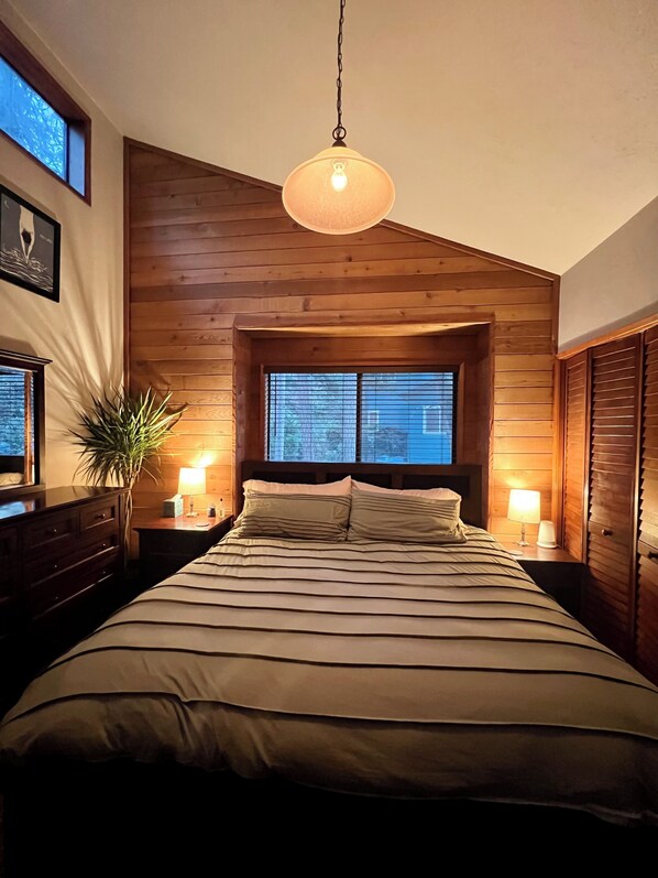 A bedroom with a large bed, wooden walls, a hanging light fixture, a window with blinds, and a bedside table with a lamp.