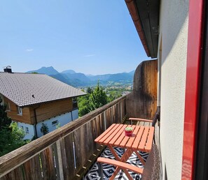 Un balcón con baranda de madera, una pequeña mesa con planta en maceta y vista a montañas y un pueblo abajo.