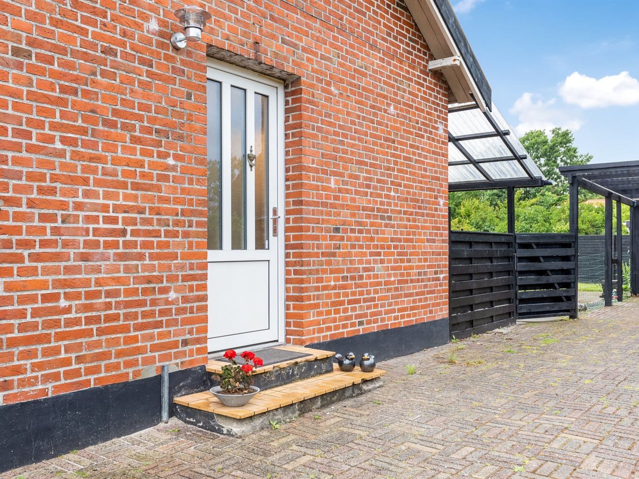 Un bâtiment en briques avec une porte blanche, un petit porche avec des marches et un jardin agrémenté de plantes en pot.