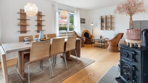 A modern living room with a dining area, wooden furniture, a black stove, and a view through a window.