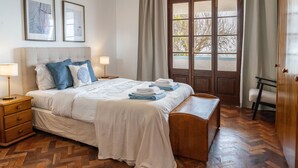 A neatly arranged bedroom with a bed, bedside tables, a bench, a window with curtains, and framed pictures on the wall. - Sailor House - By Wehost (Porto Santo)