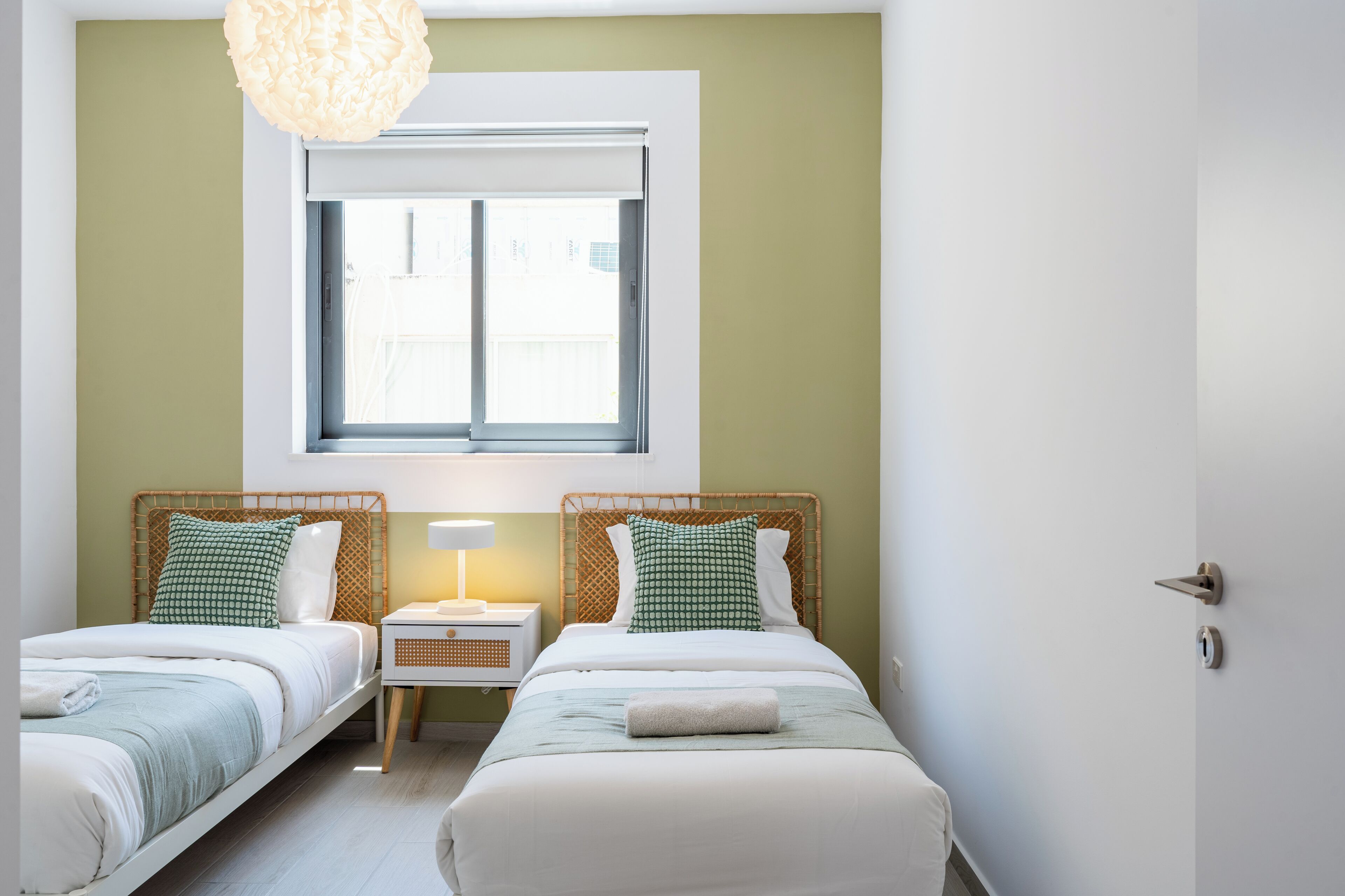 A room with two beds, a window, a bedside table with a lamp, and a hanging light fixture.