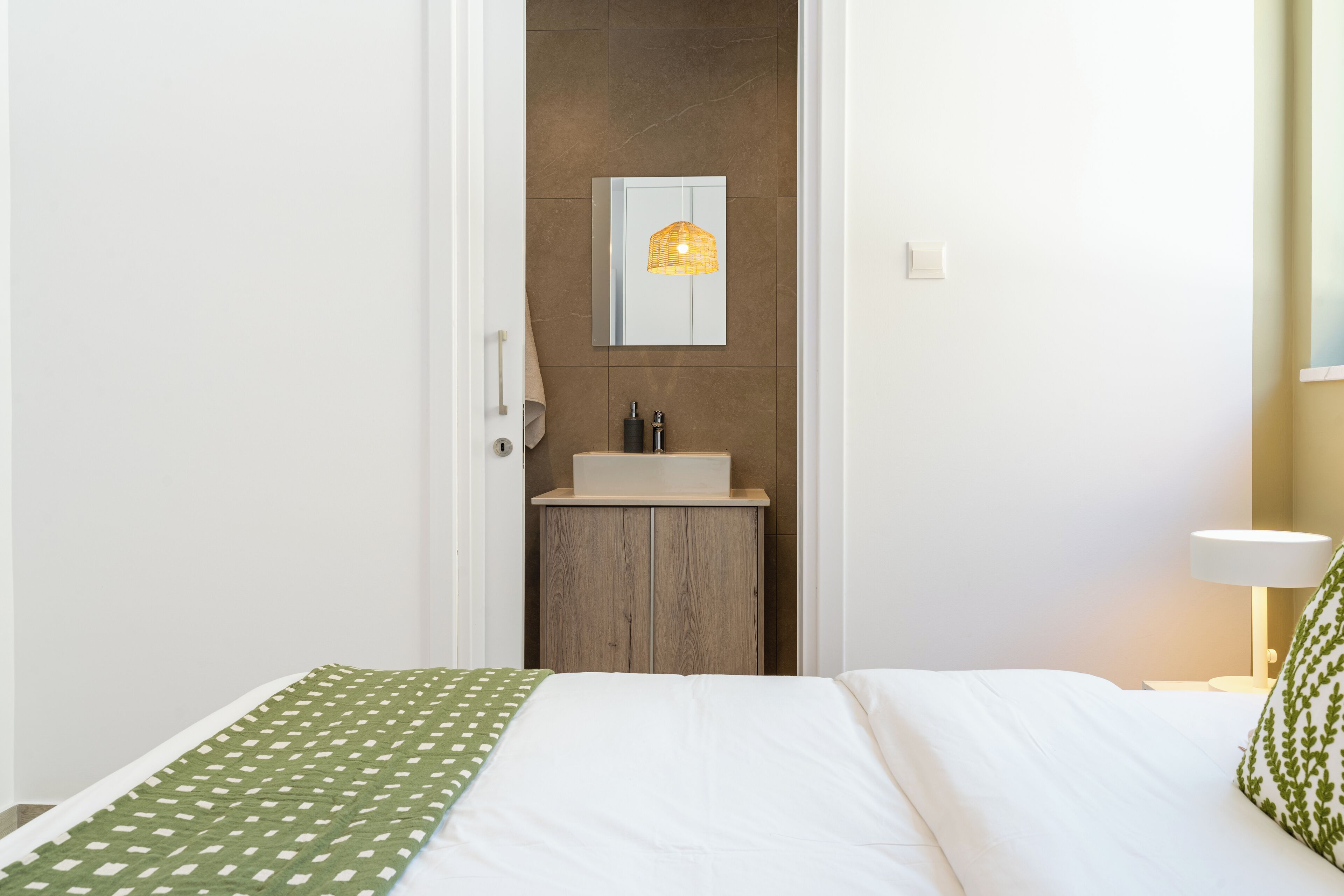 A hotel room with a bed, a small wooden cabinet with a mirror, a wall-mounted light fixture, and a bedside table with a lamp.