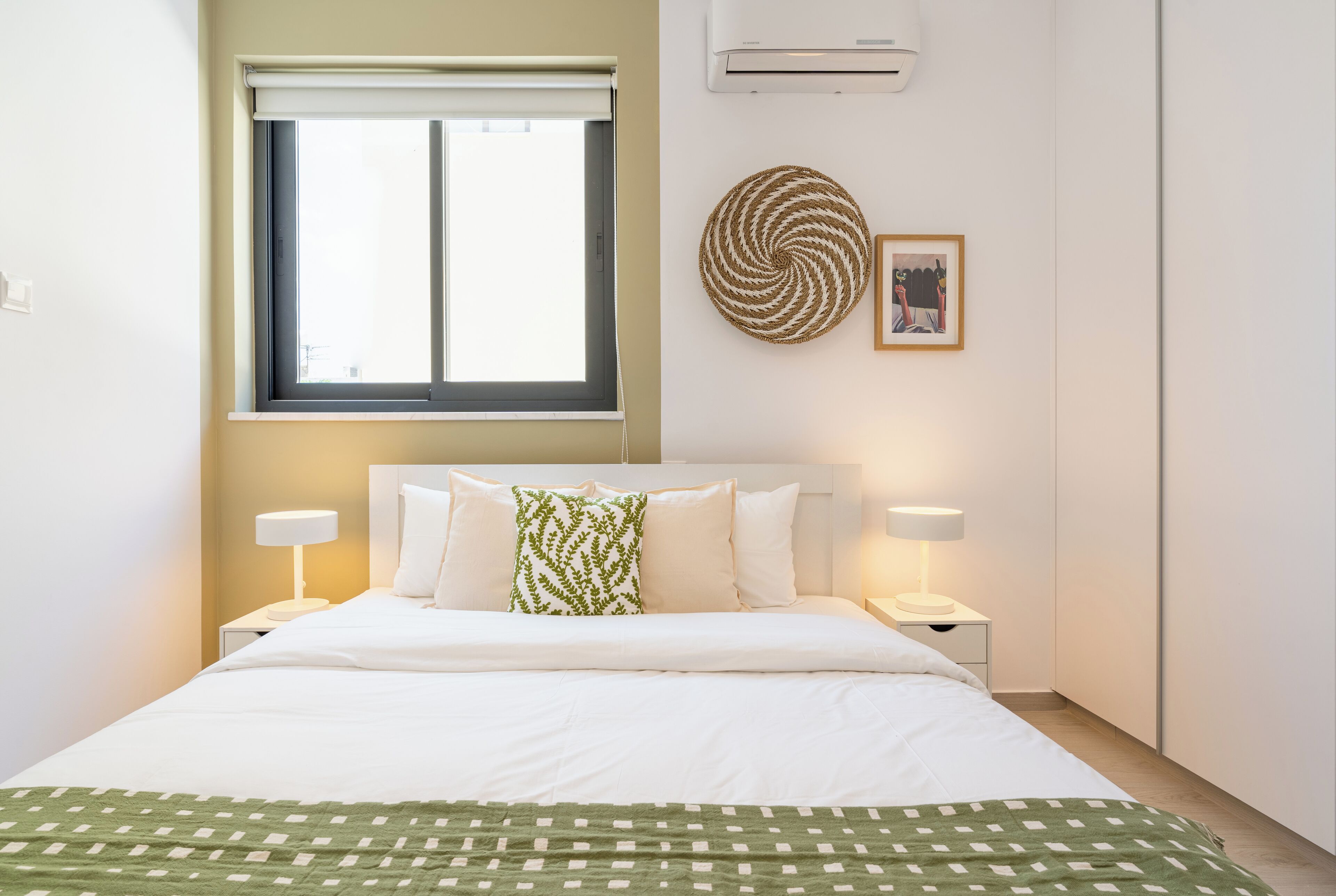 Habitación de hotel impecable con cama, dos mesitas de noche con lámparas, ventana con persiana, equipo de aire acondicionado y un adorno en la pared.