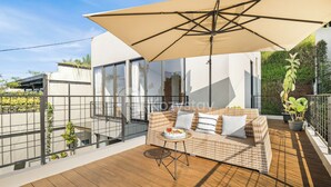 Een modern terras buiten met een rieten bank, een ronde tafel en een grote parasol.