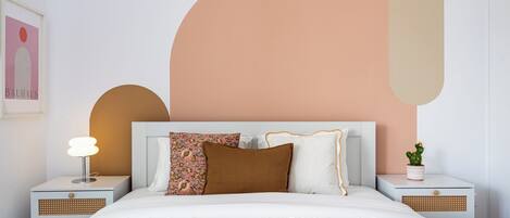 Ein ordentlich bezogenes Bett mit weißer Bettdecke, einem braunen Kissen und einem Zierkissen mit Blumenmuster. Auf dem Nachttisch links eine Lampe, rechts eine Topfpflanze.