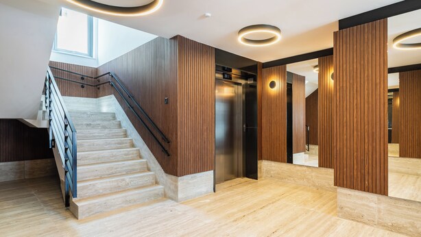 Un interior moderno con una escalera de mármol, paneles de madera y un ascensor central.