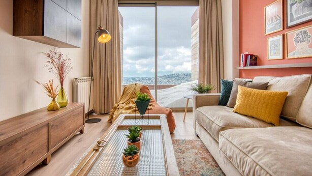 A modern living room with a sofa, a coffee table with plants, a wooden cabinet, and a view of the city through large windows. - RedRock Faqra - Mountain Getaway Residences W Pool & Balconies (Bqaatouta)